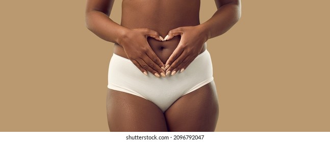 Plus Size Black Woman In Underwear Holding Hands On Abdominal Area Forming Heart Shape Around Navel, Isolated On Solid Brown Color Background, Cropped Shot. Self Love, Care, Health, Pregnancy Concept