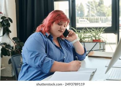 A plus size businesswoman with pink hair takes notes while on a phone call in a bright office space. - Powered by Shutterstock