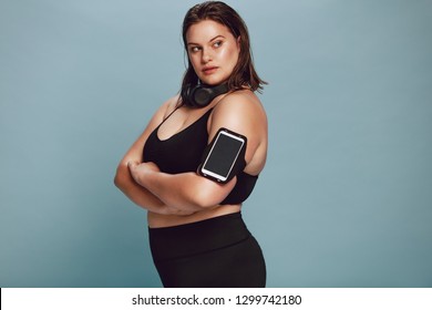 Plus Size Female Model In Sportswear With Her Arms Crossed And Looking Away. Confident Fitness Woman Posing On Grey Background With Mobile Phone On Armband And Headphones.