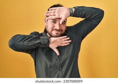 Plus size hispanic man with beard standing over yellow background smiling cheerful playing peek a boo with hands showing face. surprised and exited 