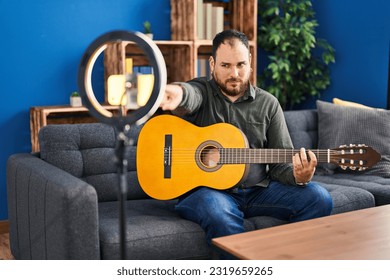 Plus size hispanic man with beard playing classic guitar at music studio recording himself pointing with finger to the camera and to you, confident gesture looking serious 