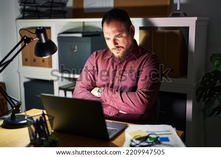 Plus size hispanic man with beard working at the office at night skeptic and nervous, disapproving expression on face with crossed arms. negative person. 