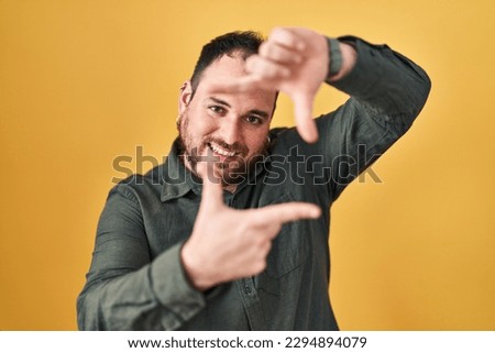Plus size hispanic man with beard standing over yellow background smiling making frame with hands and fingers with happy face. creativity and photography concept. 