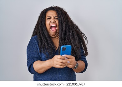 Plus Size Hispanic Woman Using Smartphone Typing Message Angry And Mad Screaming Frustrated And Furious, Shouting With Anger. Rage And Aggressive Concept. 