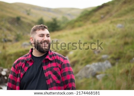 Plus size male wearing a red and black checker shirt staring off and smiling with a hilly green background