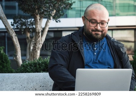 Plus size man in glasses using laptop while working outdoors. Male freelancer or IT specialist using computer for remote work
