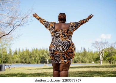Plus size model facing away from camera with arms outstretched. Park with green and blue