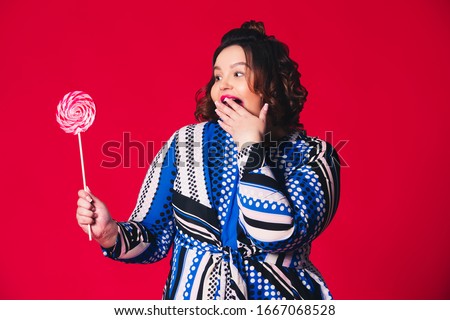 Plus size model with wide open mouth holding big lollipop on red background, body positive concept