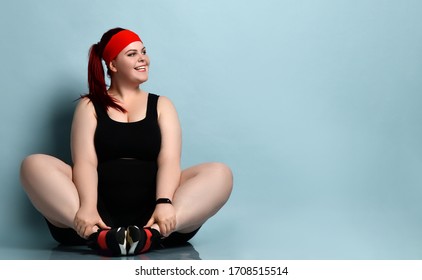 Plus Size Redhead Model In Red Headband, Black Top, Shorts, Sneakers, Fitness Bracelet. Sitting On Floor, Smiling, Looking Aside, Posing On Blue Background. Body Positive, Sport. Close Up, Copy Space