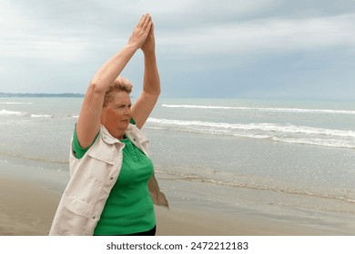 A plus size senior woman does exercises in the open air at beach against sky. Older female doing yoga to keep fit. Concept of healthy living in the elderly. Outdoors - Powered by Shutterstock