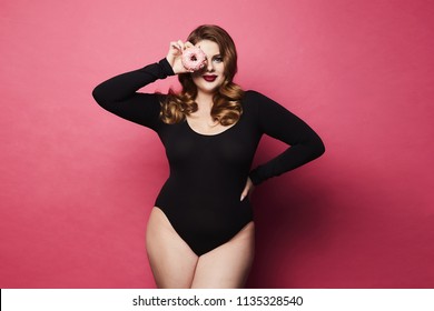 Plus size sexy model girl, fashionable blonde in black bodysuit, with bright makeup and with donut in her hand smiling and posing at pink background in studio