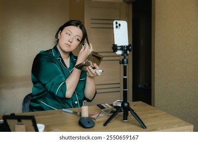 Plus size woman applying makeup on face with brush while recording vlog on smartphone for social media. Multiethnic fashion makeup artist vlogger live streaming makeup tutorial. Part of a series. UGC. - Powered by Shutterstock