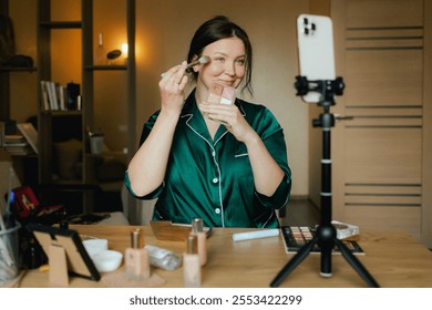 Plus size woman applying makeup on face with brush while recording vlog on smartphone for social media. Multiethnic fashion makeup artist vlogger live streaming makeup tutorial. Part of a series. UGC. - Powered by Shutterstock