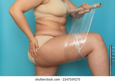 Plus Size Woman In Beige Underwear Wrap Thigh With Cling Film Closeup Side View. Body Wrapping For Weight Loss, Free Copy Space, Blue Background. Anti Cellulite Fat Burning Procedure For Slim Figure.