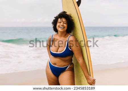 Plus size woman in a bikini holds a surfboard at the beach, ready for a thrilling surfing adventure. Female surfer smiling at the camera, reflecting the excitement of water sports and a beach vacation