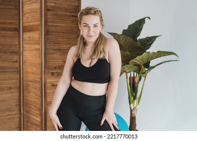 A Plus Size Woman In A Black Sports Top And Leggings. Chubby Blonde Is Engaged In Fitness. Curvy Girl Posing  In A Bright Room 