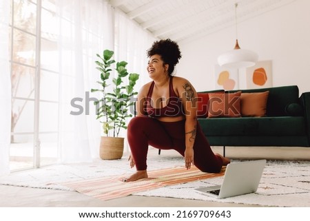 Plus size woman following workout video on laptop and doing home workout. Happy female in workout wear stretching at home.