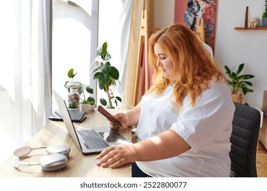 A plus size woman sits at her desk, absorbed in her phone, surrounded by a warm, inviting space. - Powered by Shutterstock