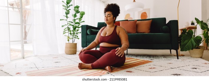 Plus size woman sitting cross legged and closed eyes in living room. Pretty female in fitness wear practicing meditation at home. - Shutterstock ID 2119985081