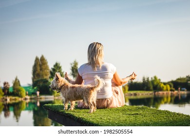 Plus Size Woman Sitting In Lotus Position And Her Cairn Terrier Dog. Rear View Of Overweight Woman Practicing Yoga On Pier At Lake. Leisure Activity Outdoors