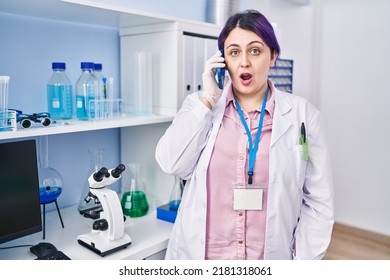 Plus size woman wit purple hair working at scientist laboratory speaking on g the phone scared and amazed with open mouth for surprise, disbelief face 