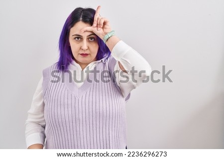 Plus size woman wit purple hair standing over white background making fun of people with fingers on forehead doing loser gesture mocking and insulting. 