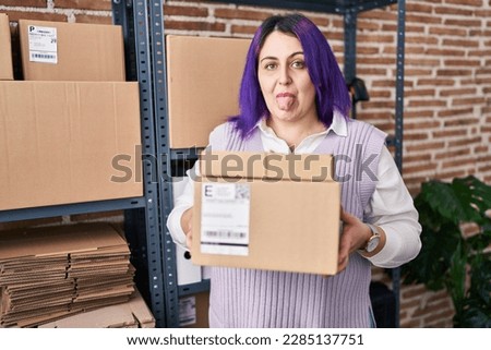 Plus size woman wit purple hair working at small business ecommerce holding boxes sticking tongue out happy with funny expression. 