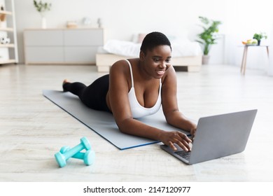 Plus Size Young Black Woman Lying On Sports Mat, Using Laptop, Speaking To Personal Coach Online Or Watching Fitness Video At Home. Overweight Millennial Lady With Pc Taking Break From Workout