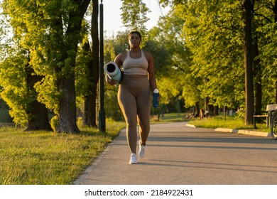 Plus Sized African American Woman Walking With Exercise Mat And Water Through The Park In A Summer Day