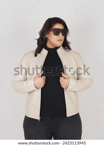 A plus sized beautiful female model posing in beige leather jacket in style with high heels and black jeans along orange bag 