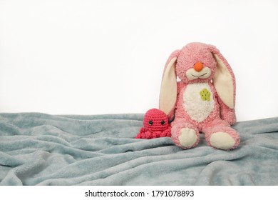 A Plush Toy Sitting On Plush Blanket And White Wall. Plush Pink Bunny Toy With Pink Knitted Octopus.