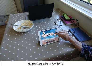 Plymouth England. July 2020. Domestic Kitchen Table With Hand Playing Solitaire On Tablet Computer. Spectacles, Open Lap Top Computer On Brown Speckled Tablecloth. Empty China Bowl With Sauce, Spoon