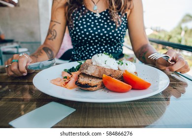 Poached Eggs Benedict Served On Toast With Smoked Salmon, Avocado, Grilled Tomato And Spinach, Young Woman Eat Breakfast With Knife And Fork