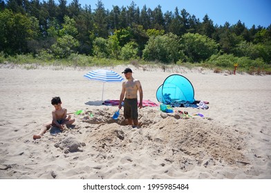 POBIEROWO, POLAND - Jun 07, 2021: Man And Child Digging A Hole In Sand Next To A Small Tent And Parasol At The Beach On A Warm Spring Day 