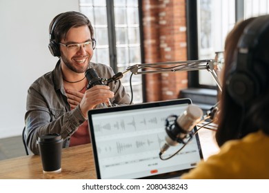 Podcast content. Excited handsome male guest gesturing, looking at female host while they making audio podcast in studio. Two podcasters in headphones laughing, talking, recording live podcast