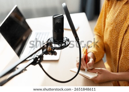 Podcaster writing notes while recording engaging audio content in a home studio, utilizing a laptop, condenser microphone, and pop filter for optimal sound quality