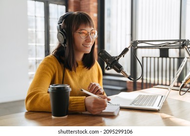 Podcasting and blogging. Young smiling woman broadcasting an audio podcast for her online show in studio, looking toward, using microphone, laptop and headphones, holding a pen, writing in notepad