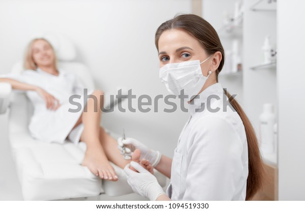 Podiatrist female doctor in mask on face, looking\
at camera and making procedure for foot with special equipment.\
Pretty blondie client spanding time at beauty salon, caring about\
her health.