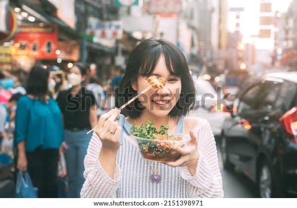 Poeple travel and eating street food\
concept. Happy young adult asian foodie woman holding spicy grilled\
squid at southeast asia Chinatown\
market.