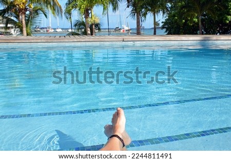 POF on woman’s crossed feet with beaded ankle bracelet relaxing by clear aquamarine pool bordered by wood deck with background of palm trees and sailboats docked on turquoise sea under clear blue sky.