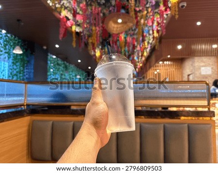 Point of view of a hand holding an empty frosted plastic cup with a straw against a blurred modern cafe background.