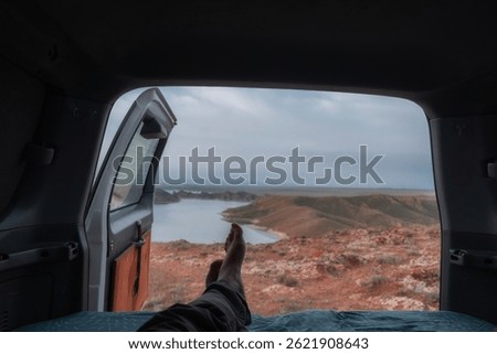 Point of view from inside a camper van looking out at Kurtinskoye Reservoir near Almaty, Kazakhstan. A person relaxes with legs stretched out, enjoying a scenic lakeside view