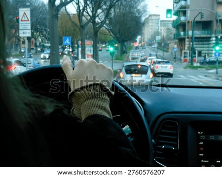 Point of view from inside the car: driver’s hand on the steering wheel while driving in city traffic. Green traffic lights, other cars ahead, urban street with trees and buildings visible through the windshield. Realistic lifestyle photography capturing daily commuting, morning drive and modern city life. High resolution interior car photo perfect for transportation, lifestyle, urban driving, business commute and advertising concepts