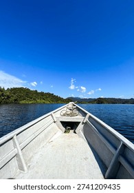 Point of view shot from the bow of a small boat moving across a calm lake surrounded by lush green tropical hills under a clear sky.