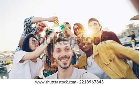 Point of view shot of handsome guy holding camera and taking selfie while his friends having fun