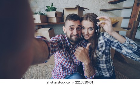 Point of view shot of happy young couple taking selfie with house keys after purchasing new apartment. Young people are holding camera posing and kissing with boxes in background. - Powered by Shutterstock