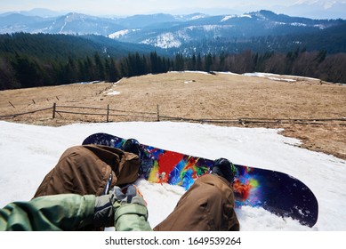 Point Of View Shot Of Snowboarder Legs And Bright Colorful Snowboard In Snow, View Of Winter Landscape. Mountain Clearing Among Tall Spruce Trees, Snowy Peaks In Background. Sports And Pov Concept.