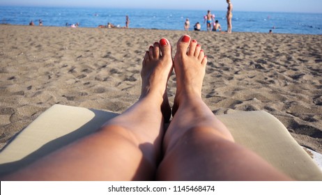 Point of view of a woman who lies on the beach and looks at her beautiful legs and the sea. Legs with red nails manicure. Calm sea, beach and people in the background in the background.