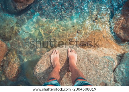 Point of view of young man with bare foot stands on turquoise water at sea beach. Mediterranean relax summer holidays.