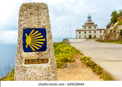 A Pointer For The Orientation Of The Camino De Santiago Pilgrims In Cape Finisterre. Finish Zero Kilometers. In The Background A Lighthouse (faro) And An Ocean. Buen Camino Peregrino